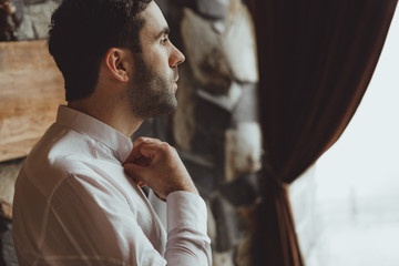 attractive man with a three-day unshaven on a beard poses as a groom before the wedding adjusting a collar on a shirt. the groom in the big building is preparing for the wedding