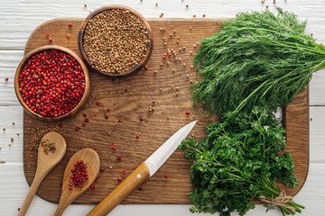 top view of green parsley and dill, knife, spoons, coriander and pink peppercorn in bowls on wooden chopping board