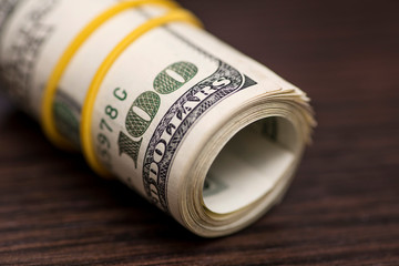 A large roll of hundred dollar bills knitted by a yellow rubber band isolated on wooden background. Roll of one hundred dollars bill on a wooden table