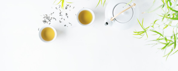 Asian tea concept, two white cups of tea and teapot surrounded with green tea dry leaves view from above, space for a text on white background.Long wide banner