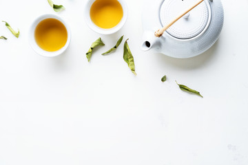 Asian tea concept, two white cups of tea and teapot surrounded with green tea dry leaves view from above, space for a text on white background.