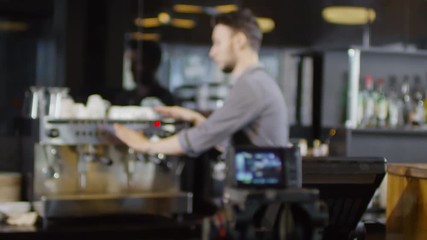 Racking focus medium shot of recording camera set on coffee shop counter, and young tattooed Caucasian barista giving demonstration of his professional coffee machine, pointing to different features