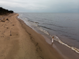 Aerial Beautiful young blonde woman beach nymph in white dress near sea with waves during a dull gloomy weather with stormy wind and rain