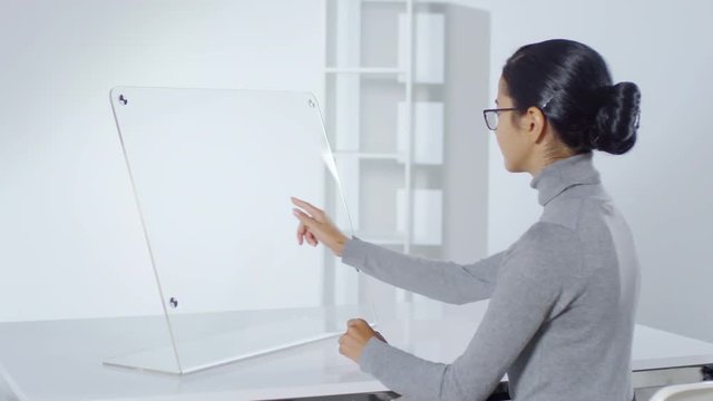 Back view of young Asian businesswoman sitting at invisible interactive screen, tapping and swiping on it. Suitable for adding augmented reality graphics