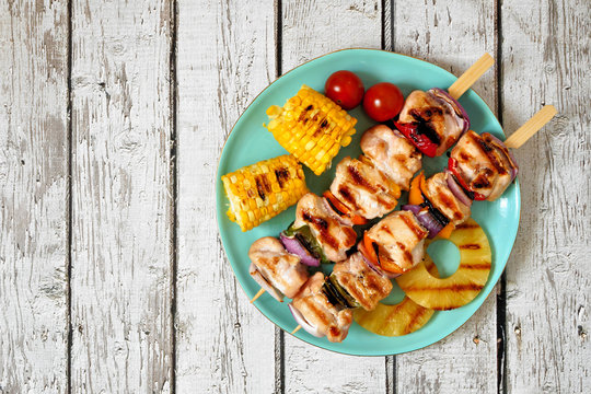 Plate Of Grilled Chicken Kabobs, Corn, And Pineapple. Top View On A White Wood Background. Summer BBQ Or Picnic Food Concept.