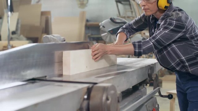 Female joiner in safety headphones and goggles pushing button of electric jointer and starting processing wooden plank in it