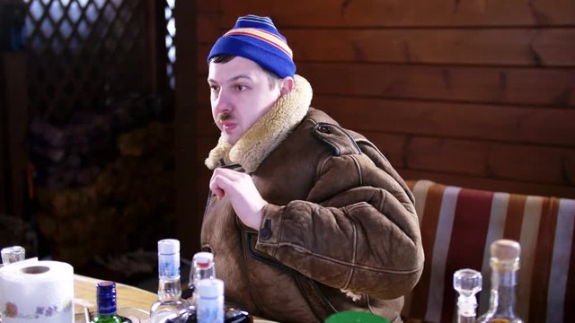 Mature caucasian funny drunk man with mustache dressed in blue cap and thick brown coat sits behind table full of food and alcoholic drinks and suddenly starts twitching outside on cold winter day.