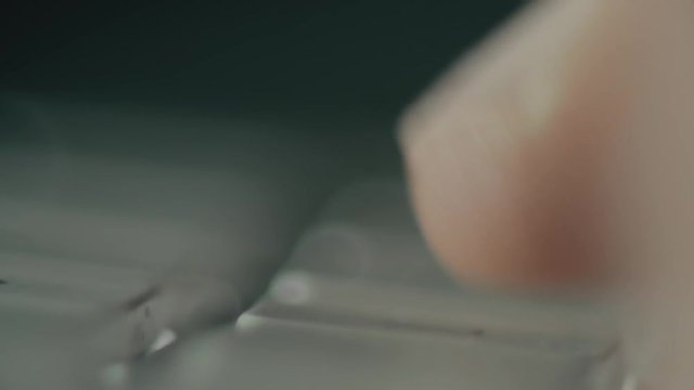 View of silver computer keyboard with white keys and fingers typing. Fingers moving over and pressing keys. Dim light shines on metallic keyboard. Extreme close up.