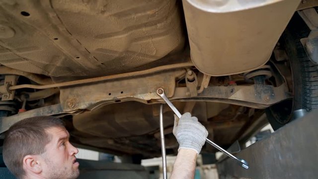 The master in the inspection pit in the service station adjusts the alignment of the car. Car lighter unscrews the nut on the car with a wrench.