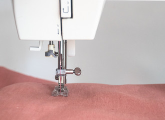Close up of foot of sewing machine of white sewing machine while working on process with pink linen fabric on white background.