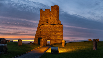 A ruin on the shore to the Irish Sea in the Welsh coastal town of Aberystwyth in the evening after sunset. The scene is lit up by many lamps.