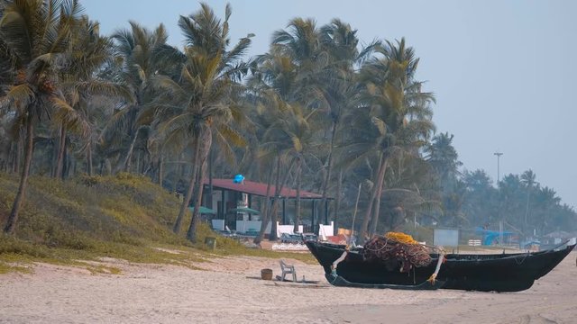 An old wooden fishing boat with a net is standing on the sand of the beach against the backdrop of palm trees, Goa, India