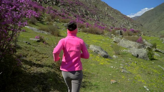 Young sporty woman running in pink alpine flowers mountain landscape. Female girl jogger jogging in nature. Athletic girl running outdoor. Healthy fitness wellness sport concept. slow-motion