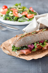 sandwich with baguette, ham, lettuce, kale on gray background, salad from kale and lettuce leaves and tomatoes on a white plate. healthy diet lunch, takeaway sandwich
