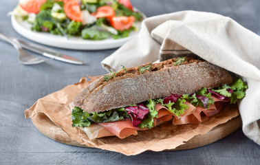 sandwich with baguette, ham, lettuce, kale on gray background, salad from kale and lettuce leaves and tomatoes on a white plate. healthy diet lunch, takeaway sandwich