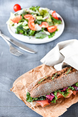 sandwich with baguette, ham, lettuce, kale on gray background, salad from kale and lettuce leaves and tomatoes on a white plate. healthy diet lunch, takeaway sandwich