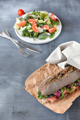 sandwich with baguette, ham, lettuce, kale on gray background, salad from kale and lettuce leaves and tomatoes on a white plate. healthy diet lunch, takeaway sandwich
