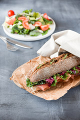 sandwich with baguette, ham, lettuce, kale on gray background, salad from kale and lettuce leaves and tomatoes on a white plate. healthy diet lunch, takeaway sandwich