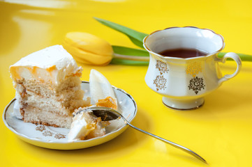A cup of tea, a plate with a piece of cake, a teaspoon and a tulip flower on a yellow background