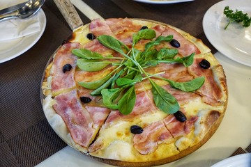 Top view of pizza with bacon, black olives topped with spinach on wooden cutting board on table in restaurant, Ready to eat or serve