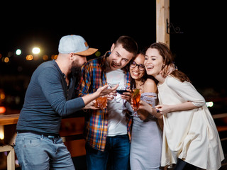 friends with smiles on their faces in the bar have fun, drink cocktails, clink glasses. friendly company celebrating on the roof against the backdrop of the night city