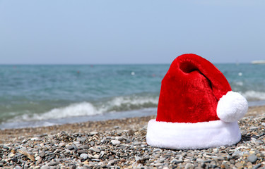 Red Santa hat on the pebbles of the beach against the backdrop of the blue sea. Horizontal, plenty of space for text, no people. Christmas concept.