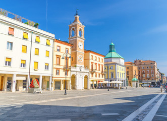 Fototapeta na wymiar Piazza Tre Martiri Three Martyrs square with traditional buildings with clock and bell tower in old historical touristic city centre Rimini with blue sky background, Emilia-Romagna, Italy