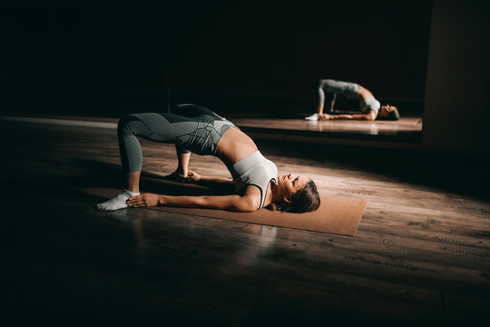 Attractive fit Caucasian woman with brown hair doing bridge on the mat. Dark gym interior, in background mirror.