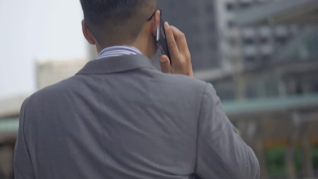 Asian businessman walking and using smartphone. Slow motion portrait asian businessman answers the phone. Asian man with glasses use phone in street near big office building.