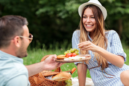 Beautiful young couple enjoying romantic picnic in a park. Relaxing on a picnic blanket and eating healthy snacks from a wooden plate