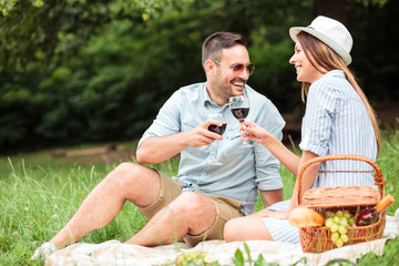 Beautiful young couple enjoying romantic picnic in a park. Relaxing on a picnic blanket and making a toast, drinking red wine.
