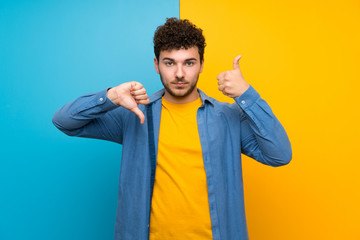 Man with curly hair over colorful wall making good-bad sign. Undecided between yes or not