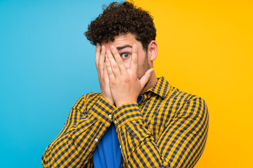 Man with curly hair over colorful wall covering eyes and looking through fingers