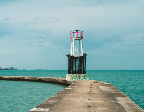 A watchtower is shown in the middle of a narrow path  in the middle of a large body of water. A cloudy day serves as the backdrop of the image.