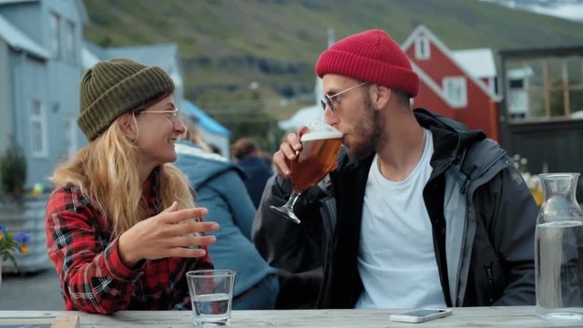 Two young millennial people from generation z, dressed in hipster outdoor gear outfits and wool beanies enjoy drink afterwork on outside terrace of cute scandinavian town, laugh and smile