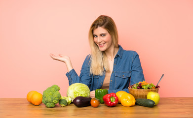 Young blonde woman with many vegetables holding copyspace imaginary on the palm to insert an ad