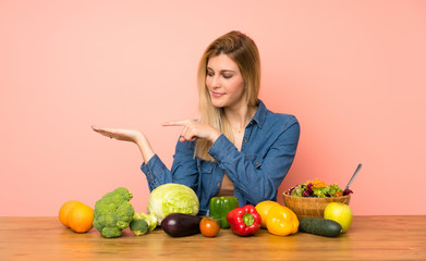 Young blonde woman with many vegetables holding copyspace imaginary on the palm to insert an ad