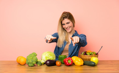 Young blonde woman with many vegetables points finger at you while smiling