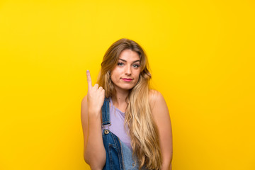 Young blonde woman with overalls over isolated yellow background pointing with the index finger a great idea
