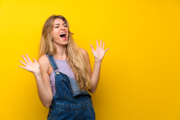 Young blonde woman with overalls over isolated yellow background with surprise facial expression