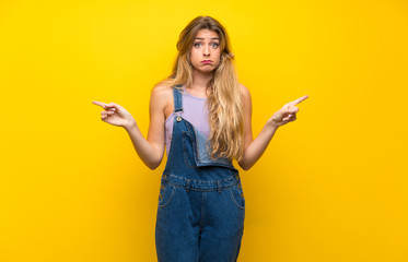 Young blonde woman with overalls over isolated yellow background pointing to the laterals having doubts