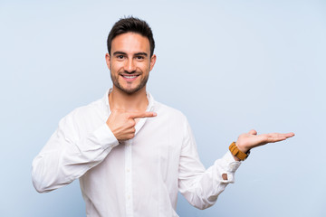 Handsome young man over isolated blue background holding copyspace imaginary on the palm to insert...