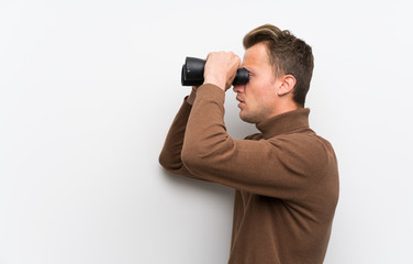 Blonde man over isolated white wall and looking in the distance with binoculars