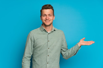Blonde man over isolated blue wall holding copyspace imaginary on the palm to insert an ad