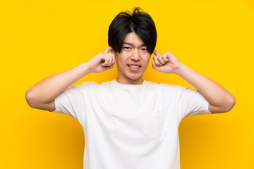 Asian man over isolated yellow wall frustrated and covering ears
