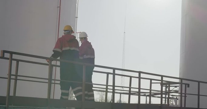 Two male industrial workers in uniform communicate at plant, fuel storage tanks on background. One uses walkie-talkie.