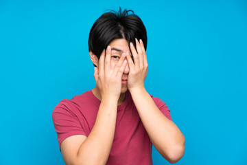 Asian man with red shirt over isolated blue wall covering eyes and looking through fingers