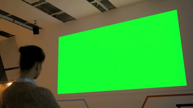 Woman looking at large blank green screen at science exhibition, museum or cinema with futuristic interior. Low light illumination. Technology, chroma key, template, mockup and entertainment concept