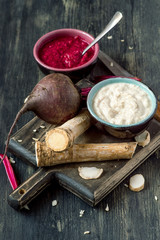 Two small round cups with freshly made seasoning, a horseradish root and beets on a dark wooden background. Selective focus.