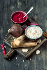 Two small round cups with freshly made seasoning, a horseradish root and beets on a dark wooden background. Selective focus.
